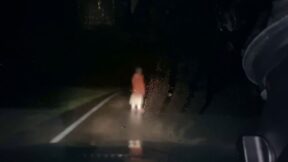 在恐怖电影“Dashcam”中，一个穿着红色外套的模糊人通过挡风玻璃在黑暗的路上出现。