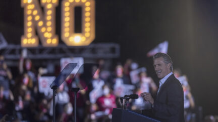 在选举集会上，加文·纽森在一个用灯泡拼成的巨大标牌前向人群发表讲话