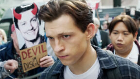 汤姆·霍兰德饰演的彼得·帕克穿过一场写着“魔鬼伪装”的抗议活动